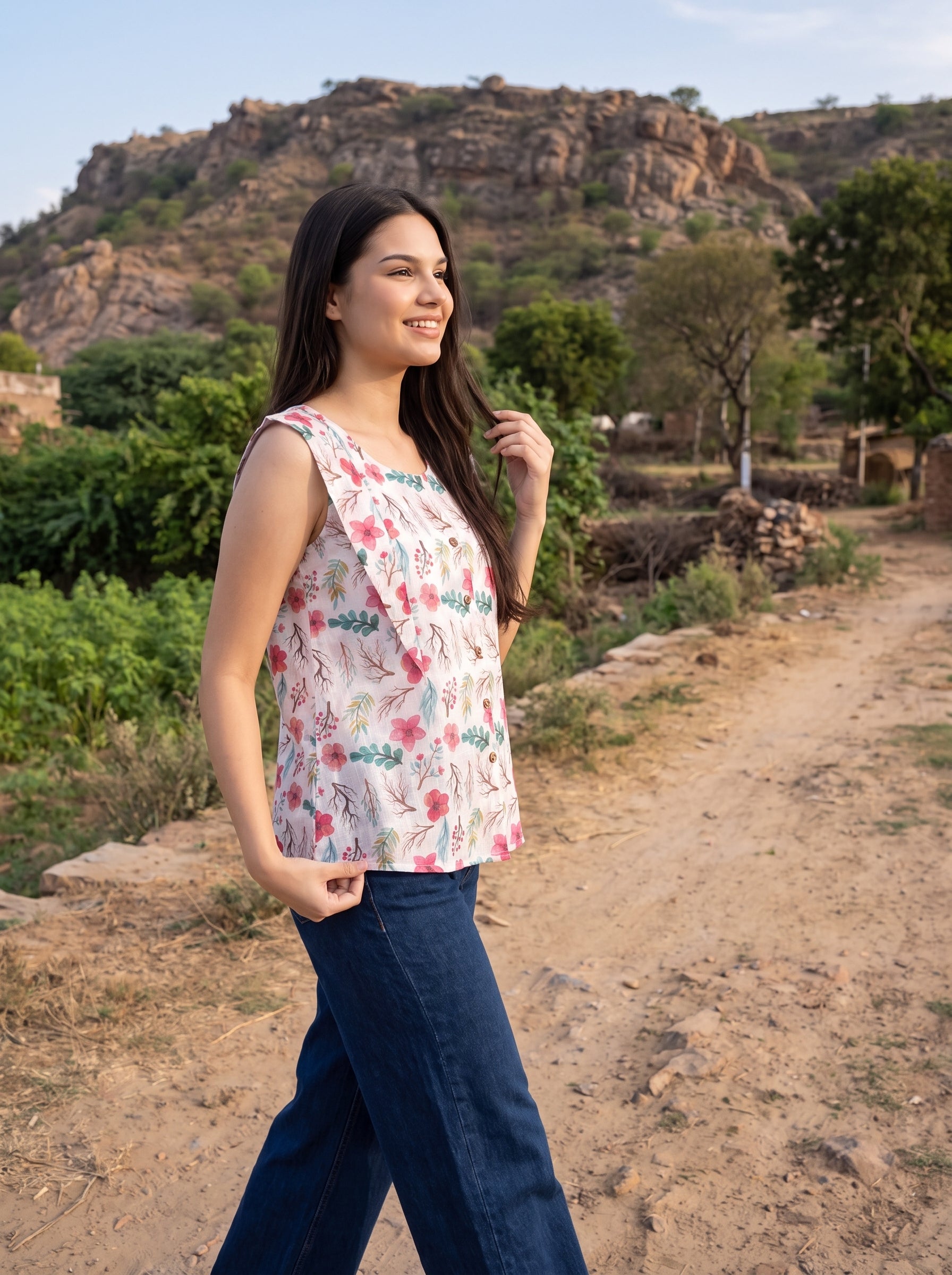 A woman wearing a sleeveless white top with a pink and green floral print and dark blue wide-leg jeans, standing on a dirt path with a rocky hill and green trees in the background.
