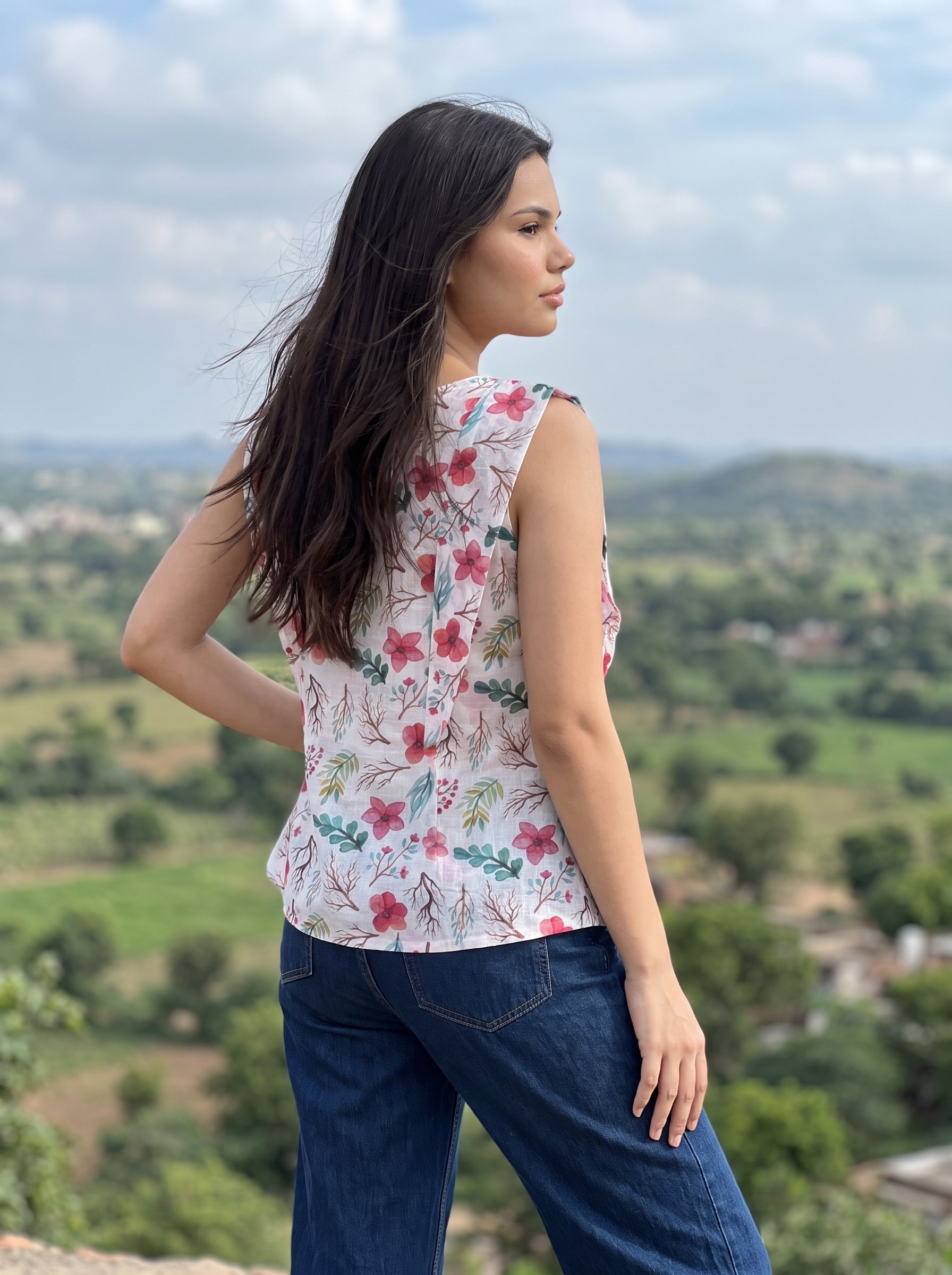 A woman wearing a sleeveless white top with a pink and green floral print, paired with dark blue jeans, standing outdoors with a scenic landscape in the background.