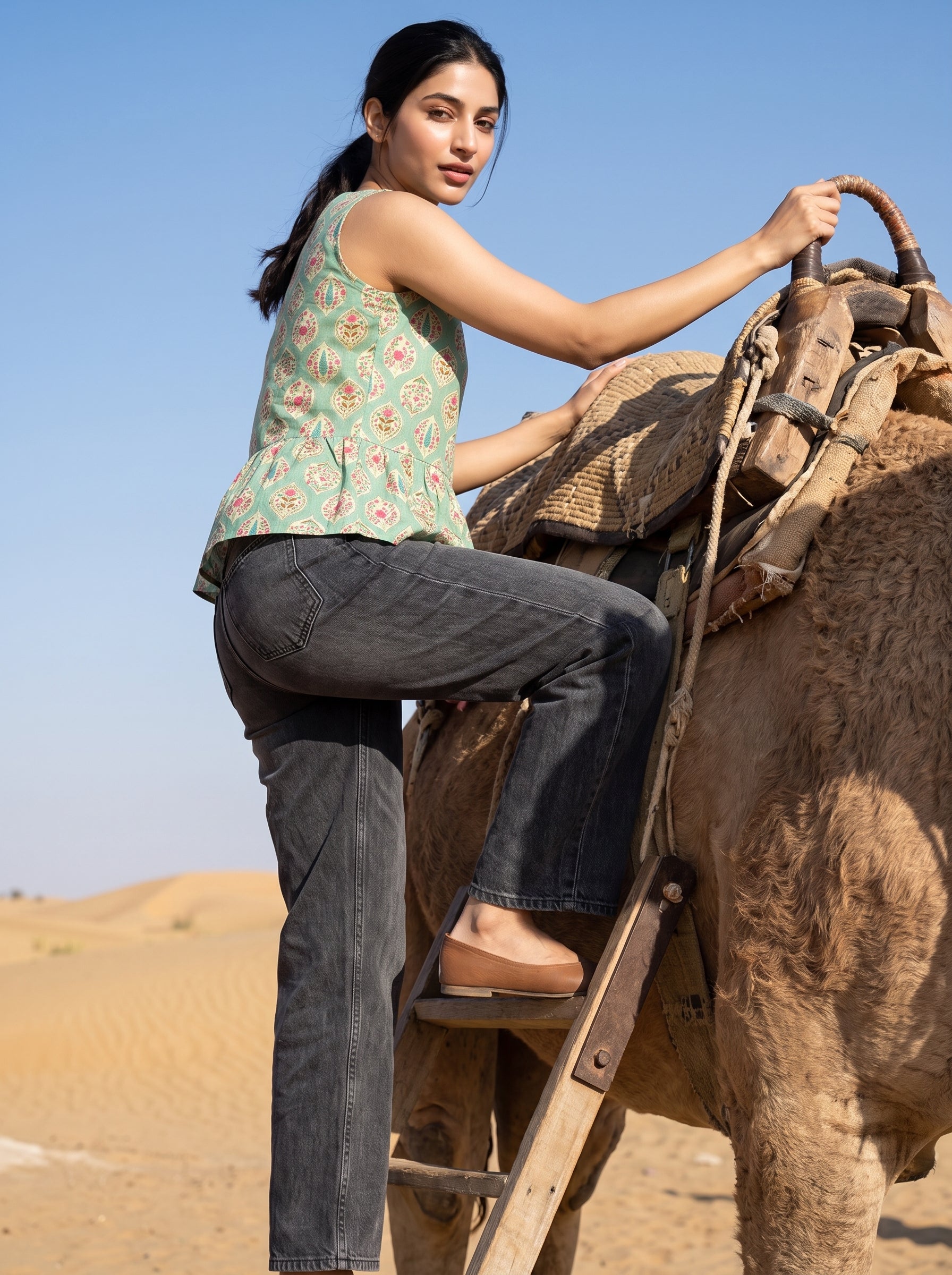 A woman in a green floral sleeveless top and dark gray jeans climbs onto a camel in a desert landscape under a clear blue sky.