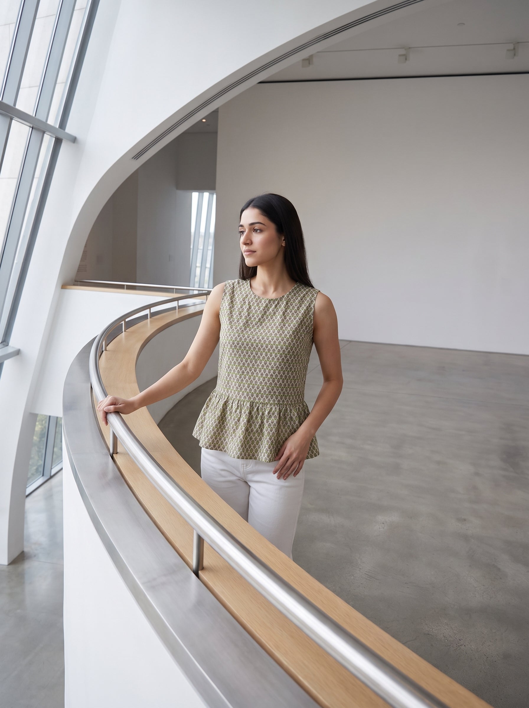 A woman stands on a curved indoor balcony wearing a sleeveless textured peplum top in a muted olive-beige tone with white pants, photographed in a modern minimalist interior with large windows and a concrete floor.