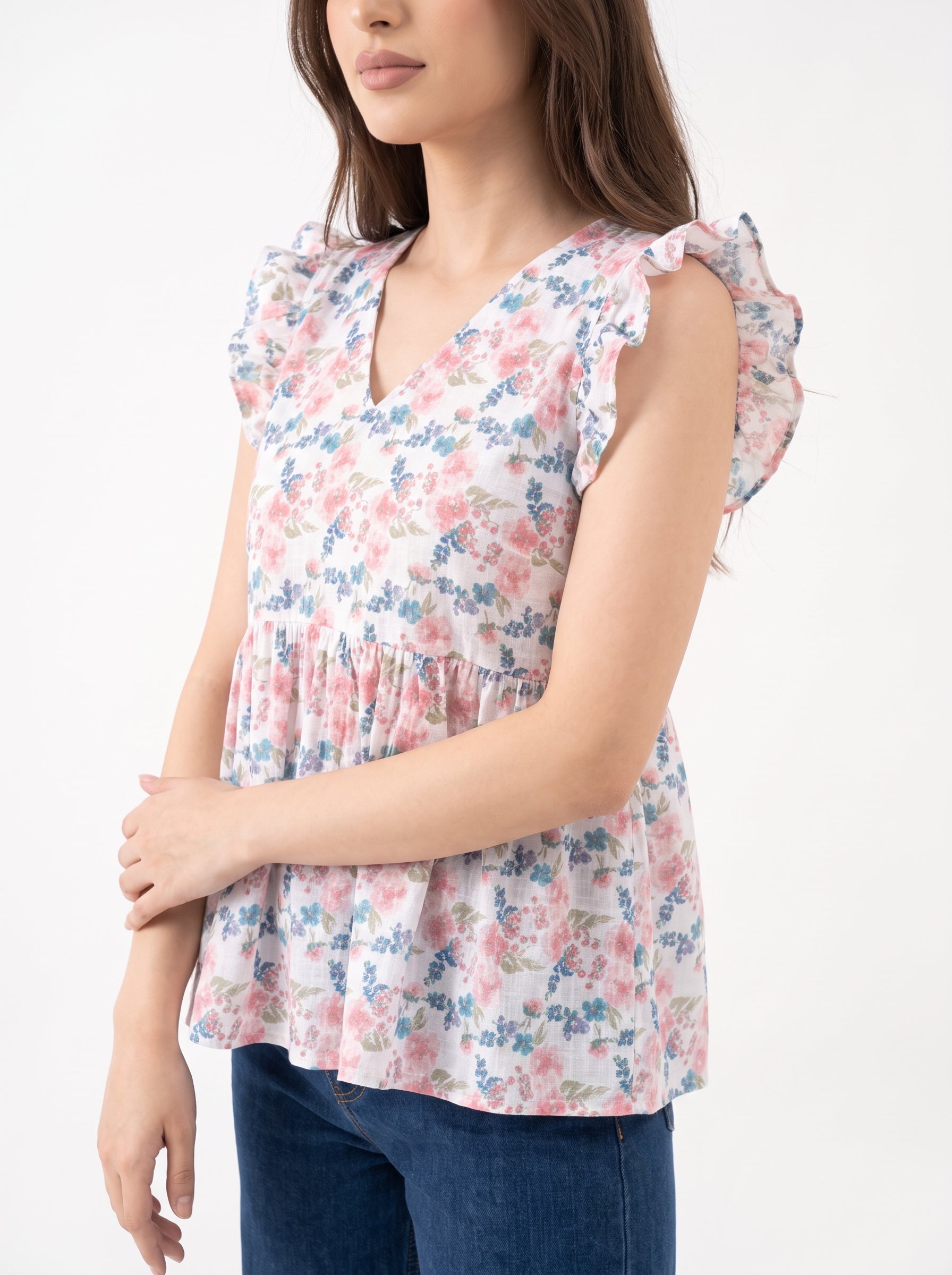 A woman wearing a sleeveless V-neck blouse with a pink, blue, and white floral print and ruffled shoulder details, paired with dark blue jeans against a plain white background.
