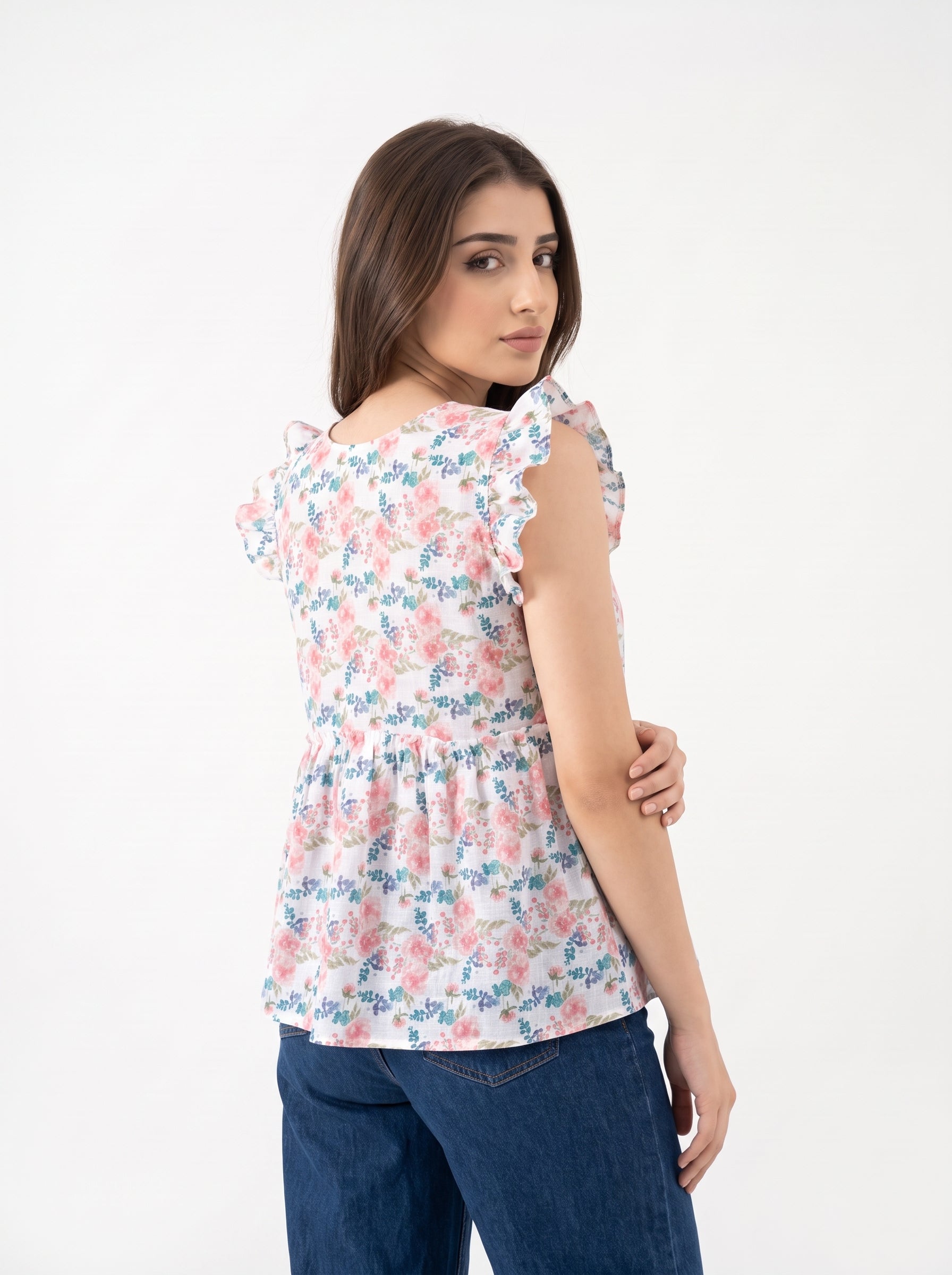 A woman wearing a white ruffled sleeveless top with a pink and blue floral print, paired with blue jeans, photographed against a plain white studio background.
