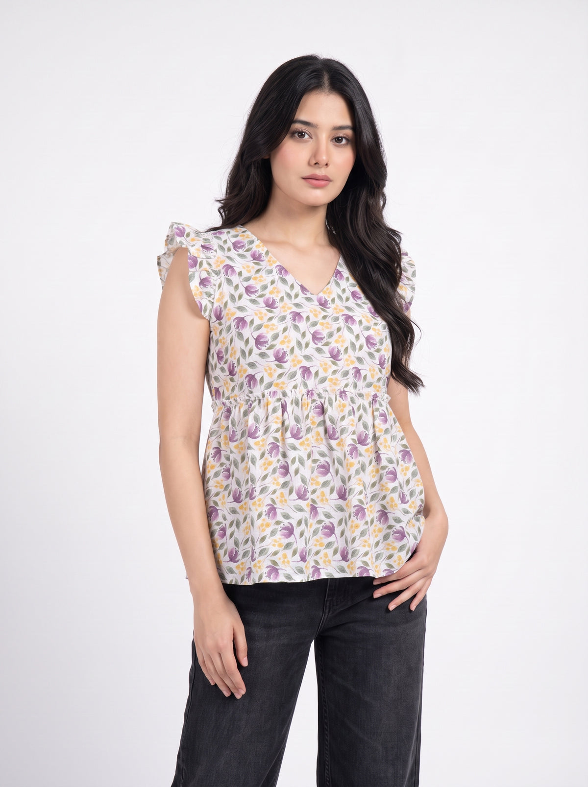 A woman modeling a sleeveless V-neck blouse with a purple and yellow floral print on a white base, paired with dark gray jeans, against a plain white studio background.