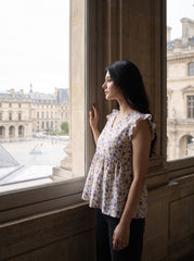 A woman with long black hair stands in profile by a large window, wearing a sleeveless peplum top with a purple and green floral print on a white base. She is looking out onto a city courtyard with classical stone buildings.