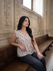 A woman with long dark hair sits on a wooden bench in a stone interior, wearing a short-sleeve V-neck blouse with a purple, yellow, and green floral pattern on a white base, paired with dark gray jeans.