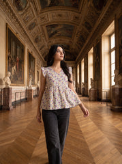 A woman wearing a short-sleeve floral peplum top and dark gray pants walks down a grand, historic gallery with wooden floors, paintings, and statues.