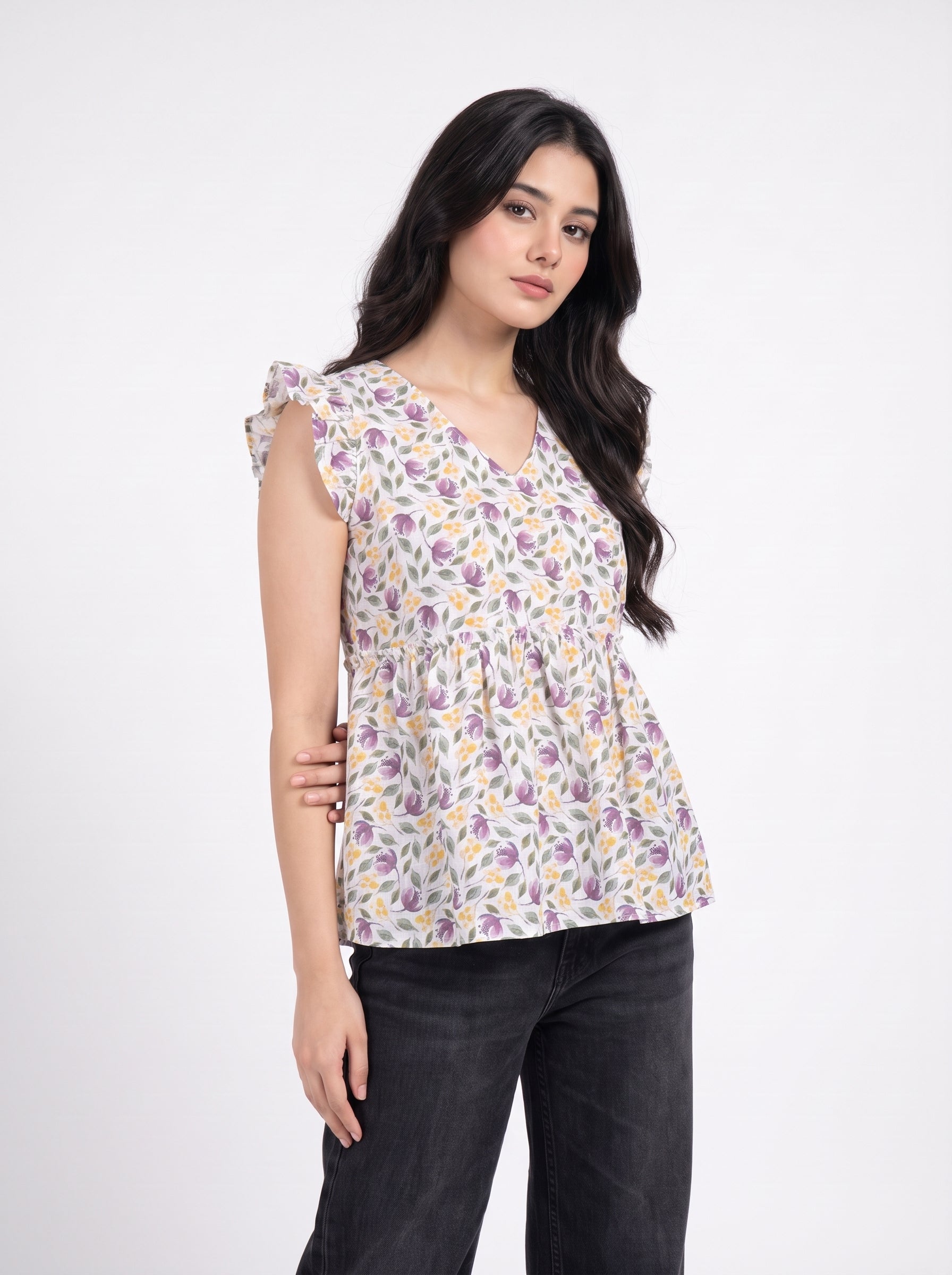 A woman modeling a sleeveless V-neck blouse with a purple and yellow floral pattern on a white base, paired with black jeans, against a plain white studio background.