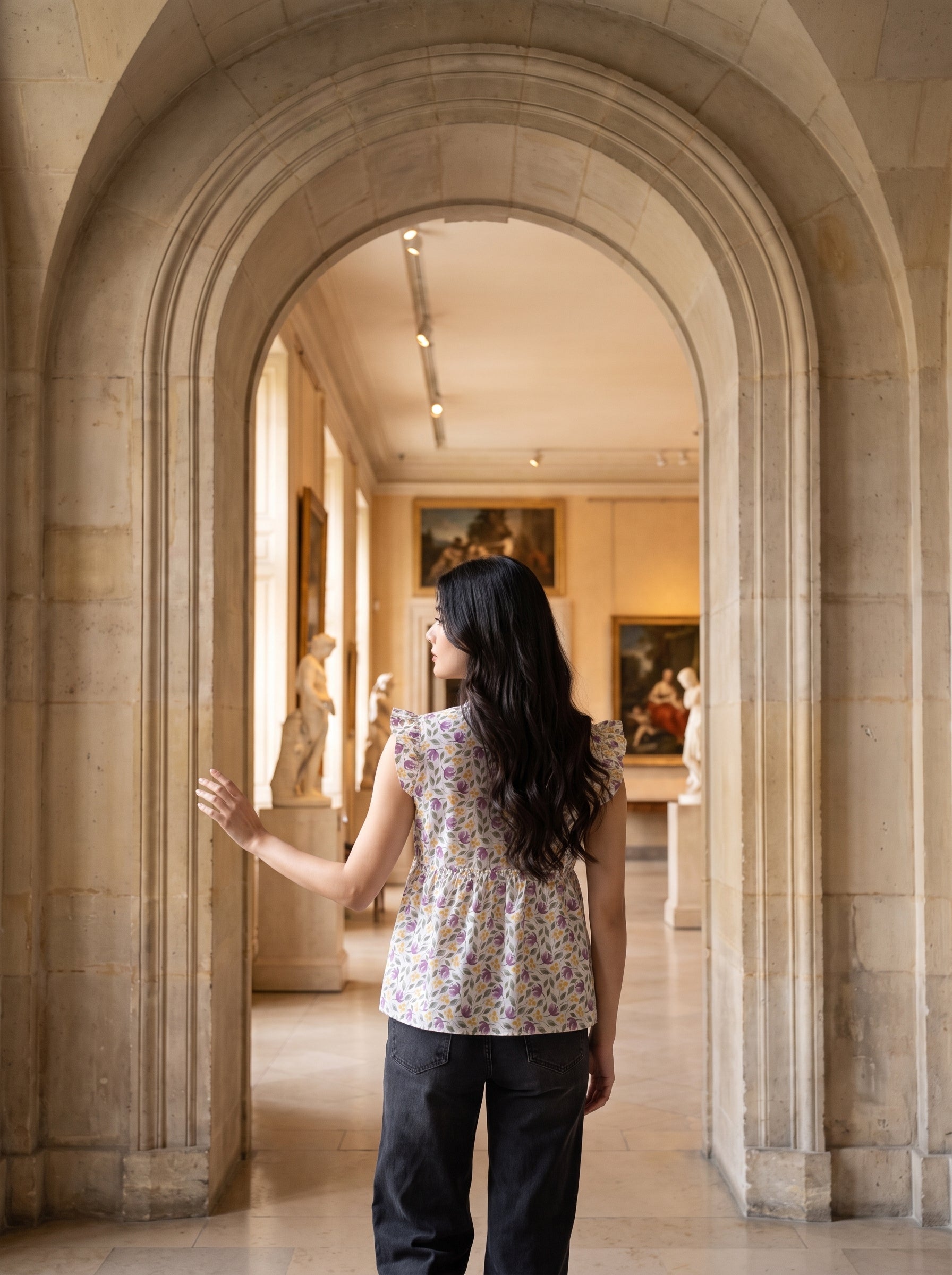 A woman with long black hair stands in a museum gallery, viewed from behind as she looks through a large stone archway. She is wearing a sleeveless floral top and dark pants.