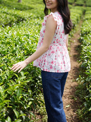 A woman standing in a green tea plantation wearing a sleeveless white top with a pink cherry blossom floral print and dark blue jeans.