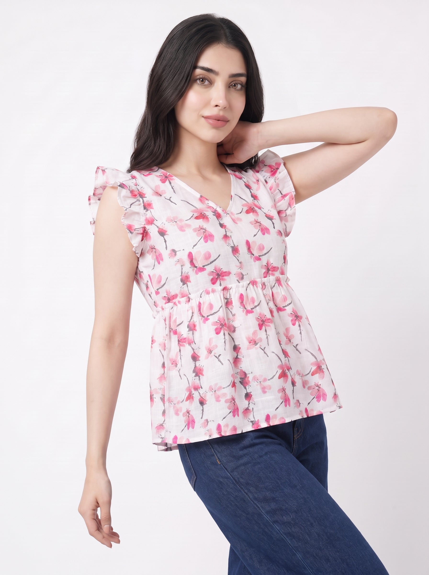 A woman modeling a white short-sleeve blouse with a pink floral print and ruffled shoulders, paired with dark blue jeans, against a plain white studio background.