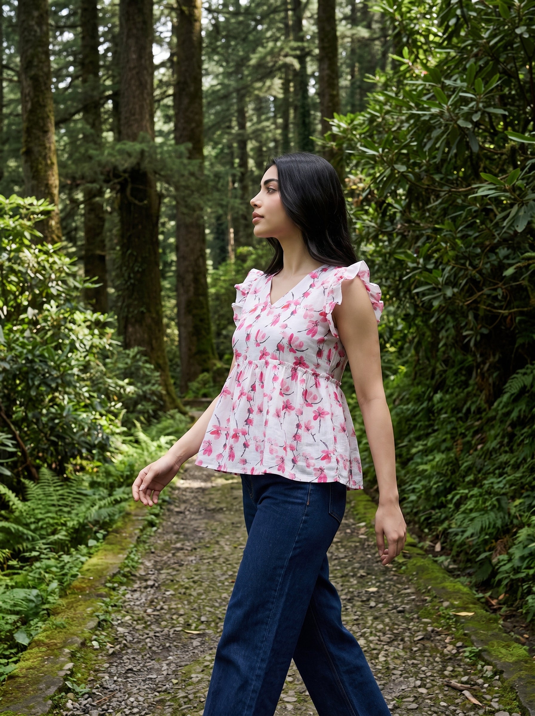 A woman wearing a pink and white floral peplum top and dark blue jeans walks along a forest path surrounded by green trees and foliage.