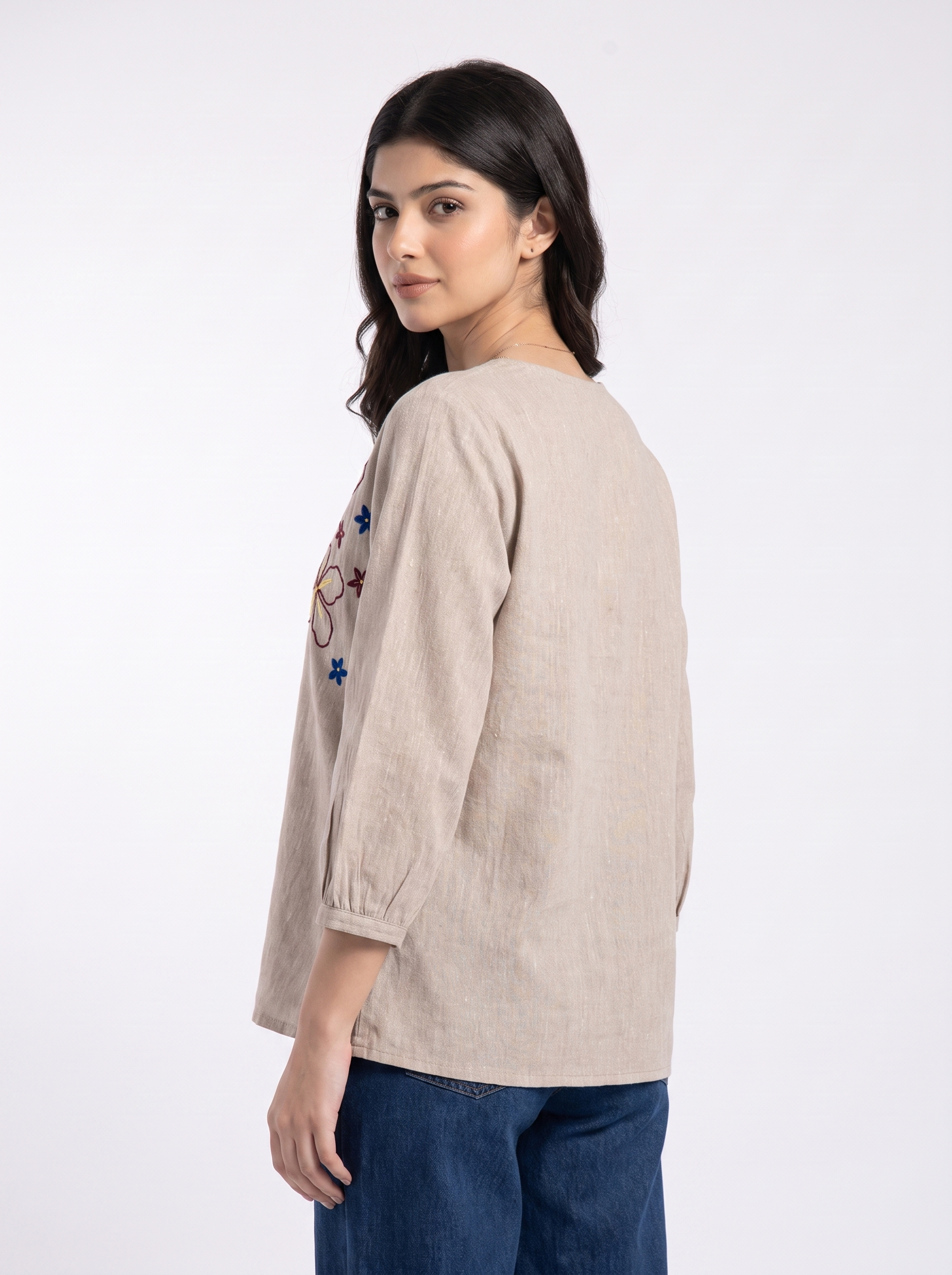 A woman wearing a beige linen long-sleeve top with colorful embroidered details on the back, paired with dark blue jeans, photographed against a plain white studio background.