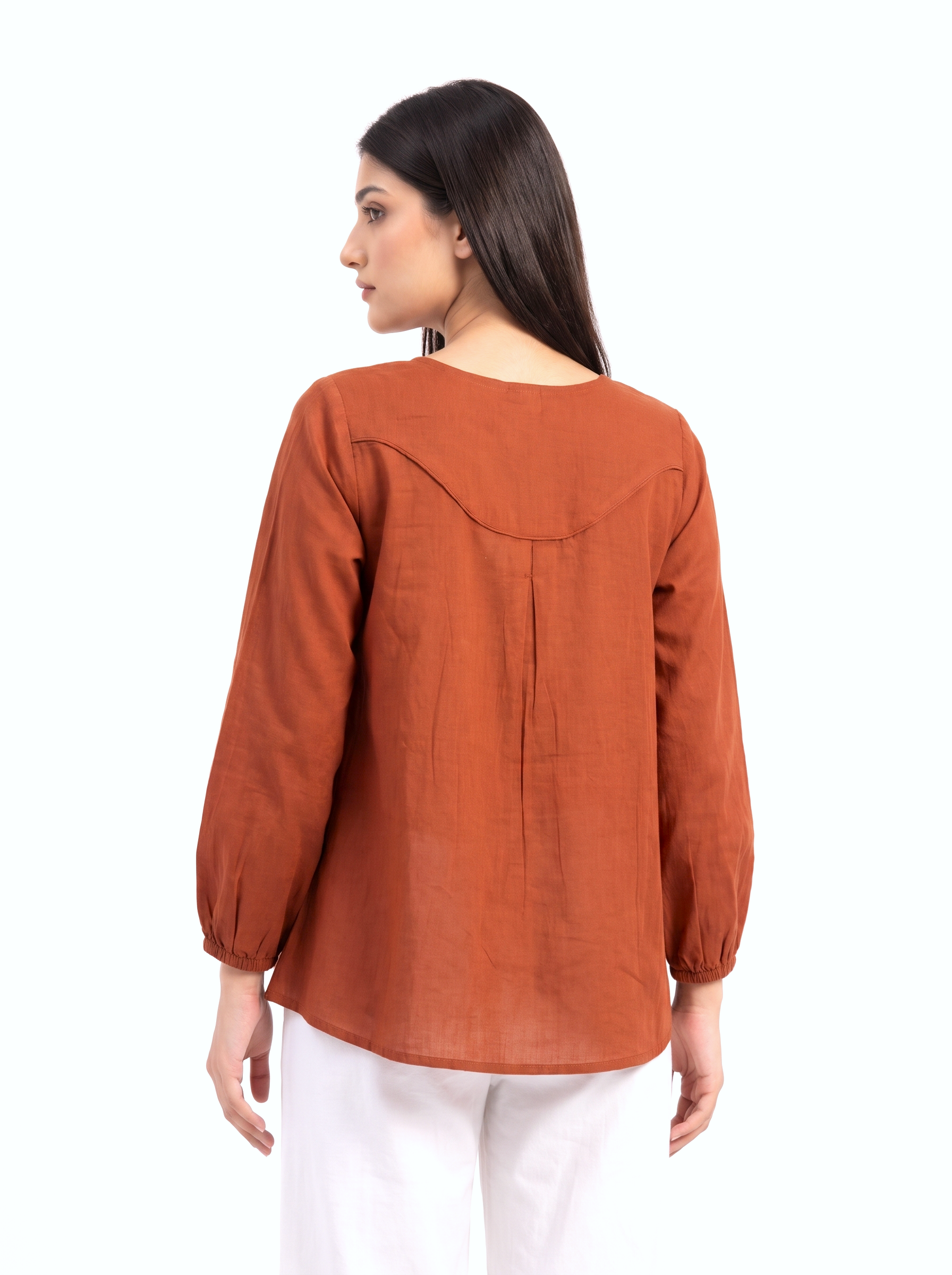A woman shown from the back wearing a rust-orange long-sleeve top with gathered sleeves and a curved seam detail across the upper back, paired with white pants against a plain white background.