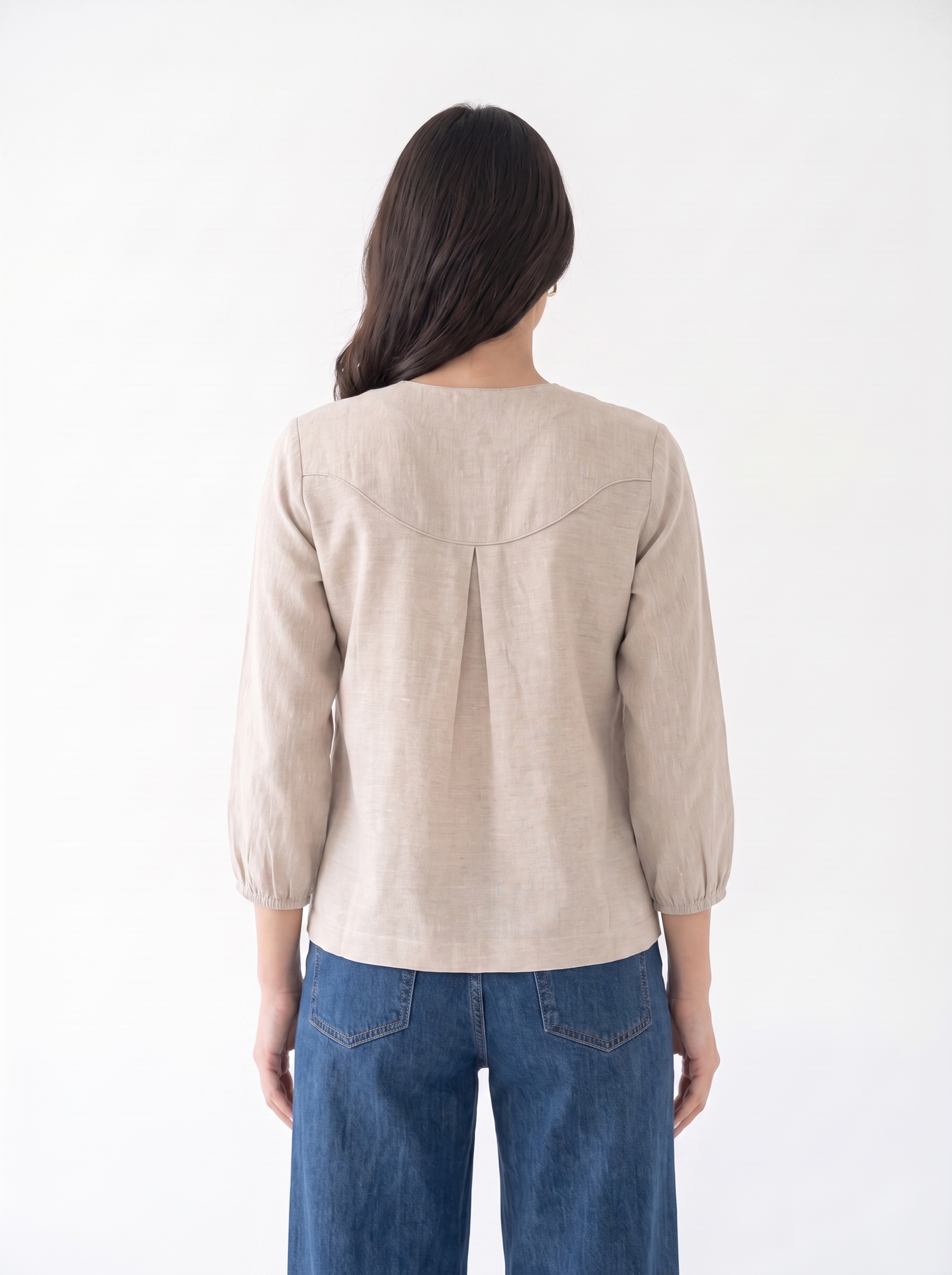 A woman viewed from the back wearing a light beige linen blouse with three-quarter sleeves and a gathered back seam, paired with blue jeans, against a plain white background.