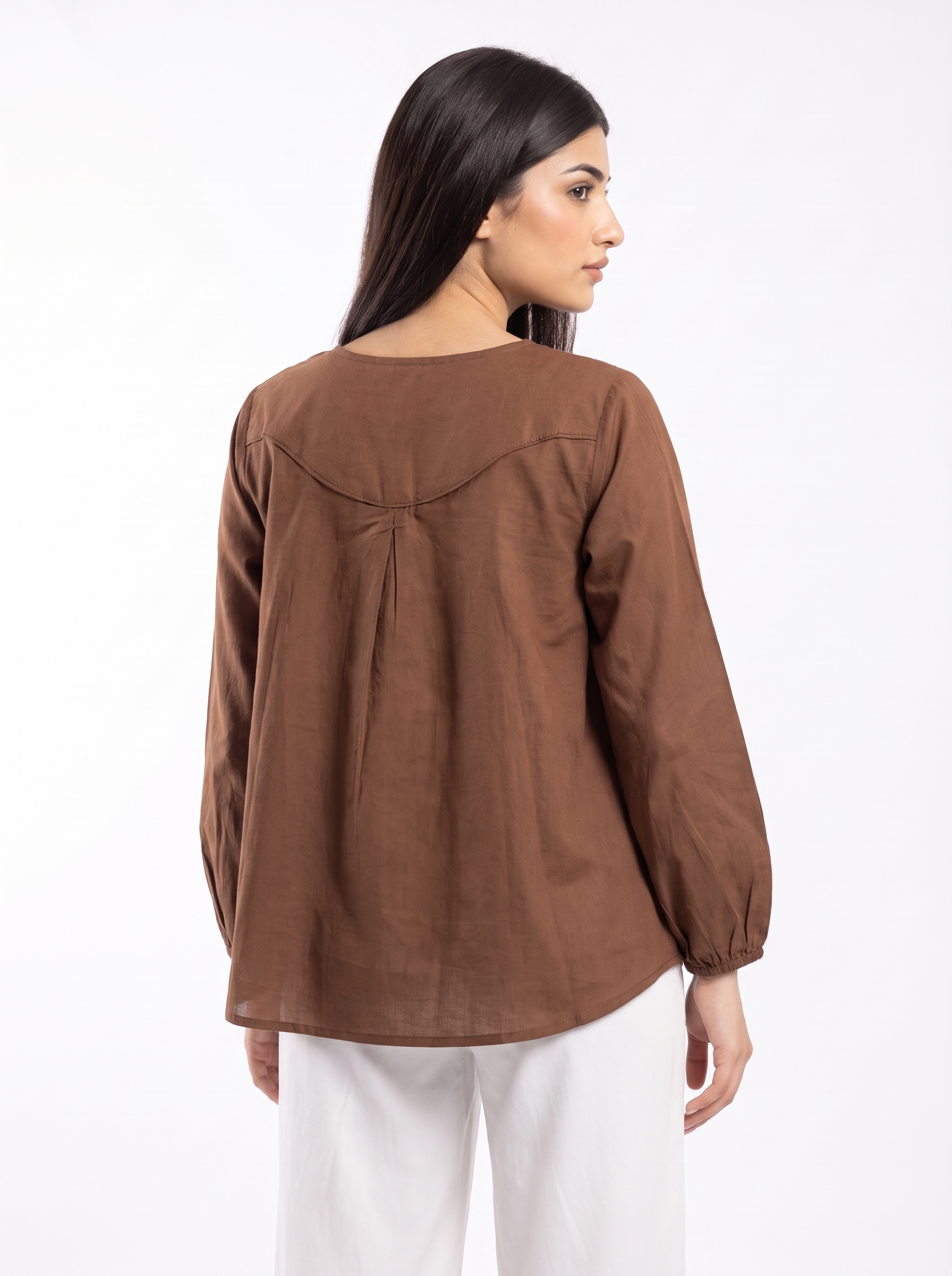 A woman shown from the back wearing a brown long-sleeve top with a gathered back detail and white pants, photographed against a plain white studio background.