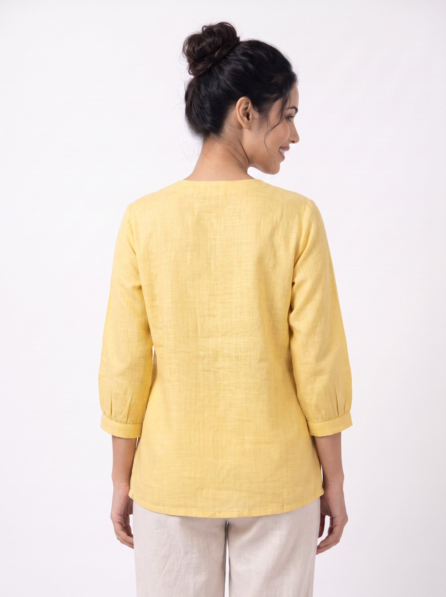 A woman shown from the back wearing a light yellow linen three-quarter sleeve top and off-white trousers, standing against a plain white studio background.