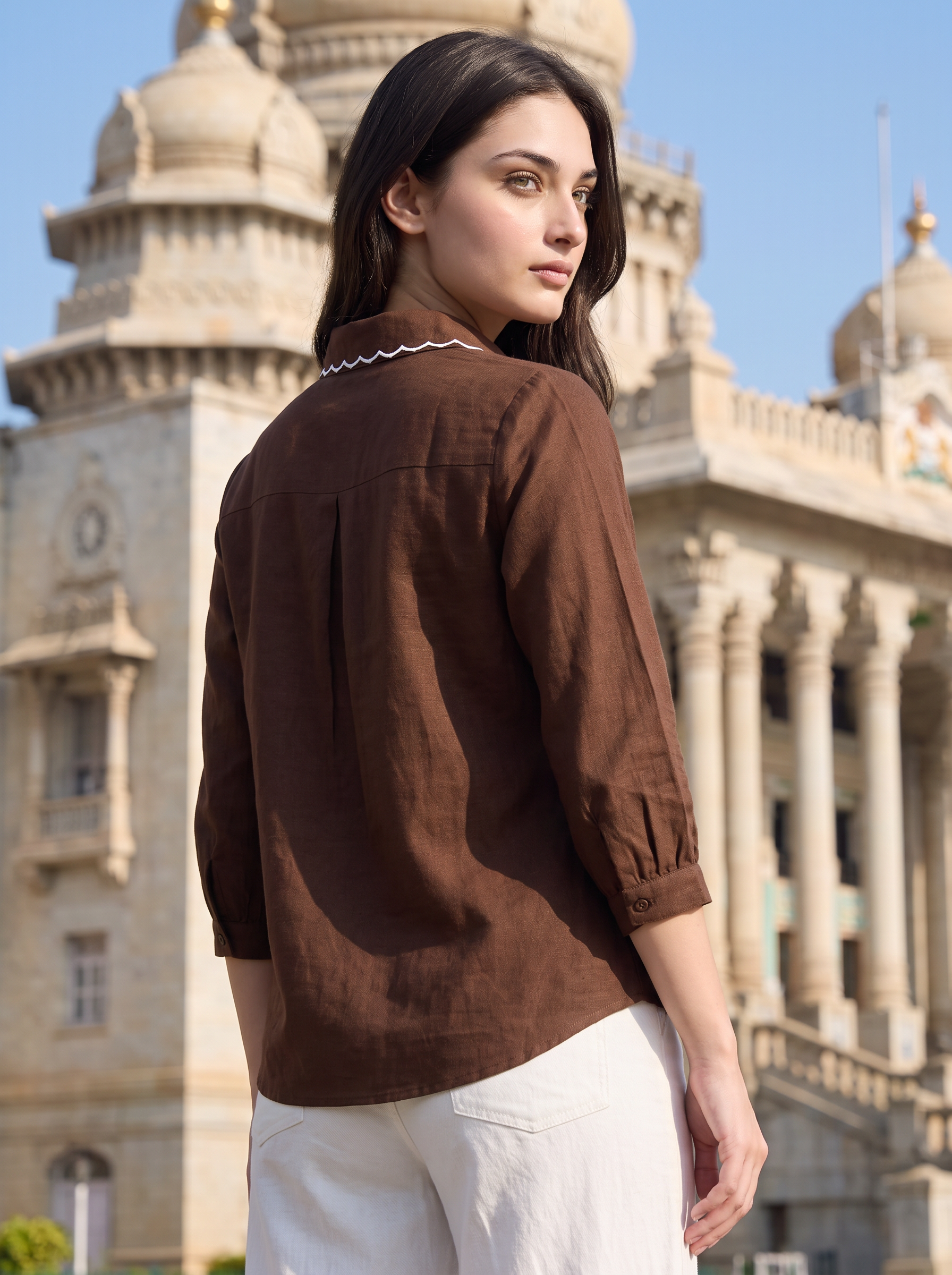 A woman wearing a brown linen three-quarter sleeve blouse with white pants, photographed outdoors in front of a grand classical building.