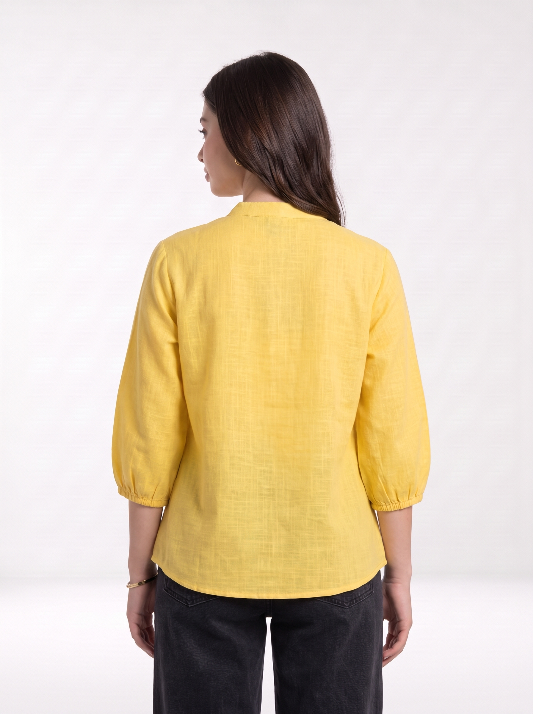 A woman viewed from the back wearing a bright yellow linen blouse with three-quarter puff sleeves and dark pants, set against a plain white studio background.