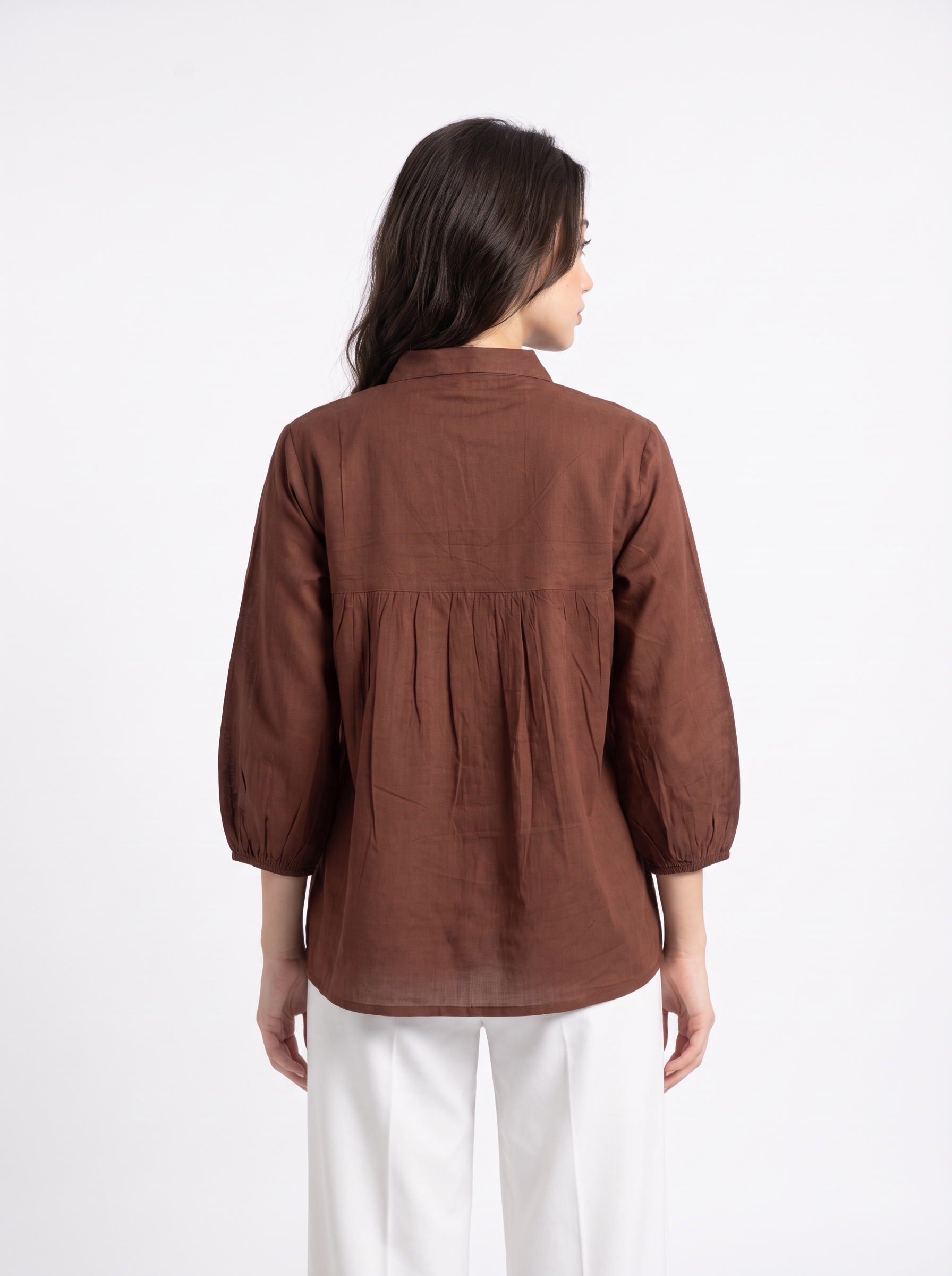 A woman viewed from the back wearing a brown long-sleeve linen blouse with a gathered waist seam and white trousers, set against a plain white studio background.