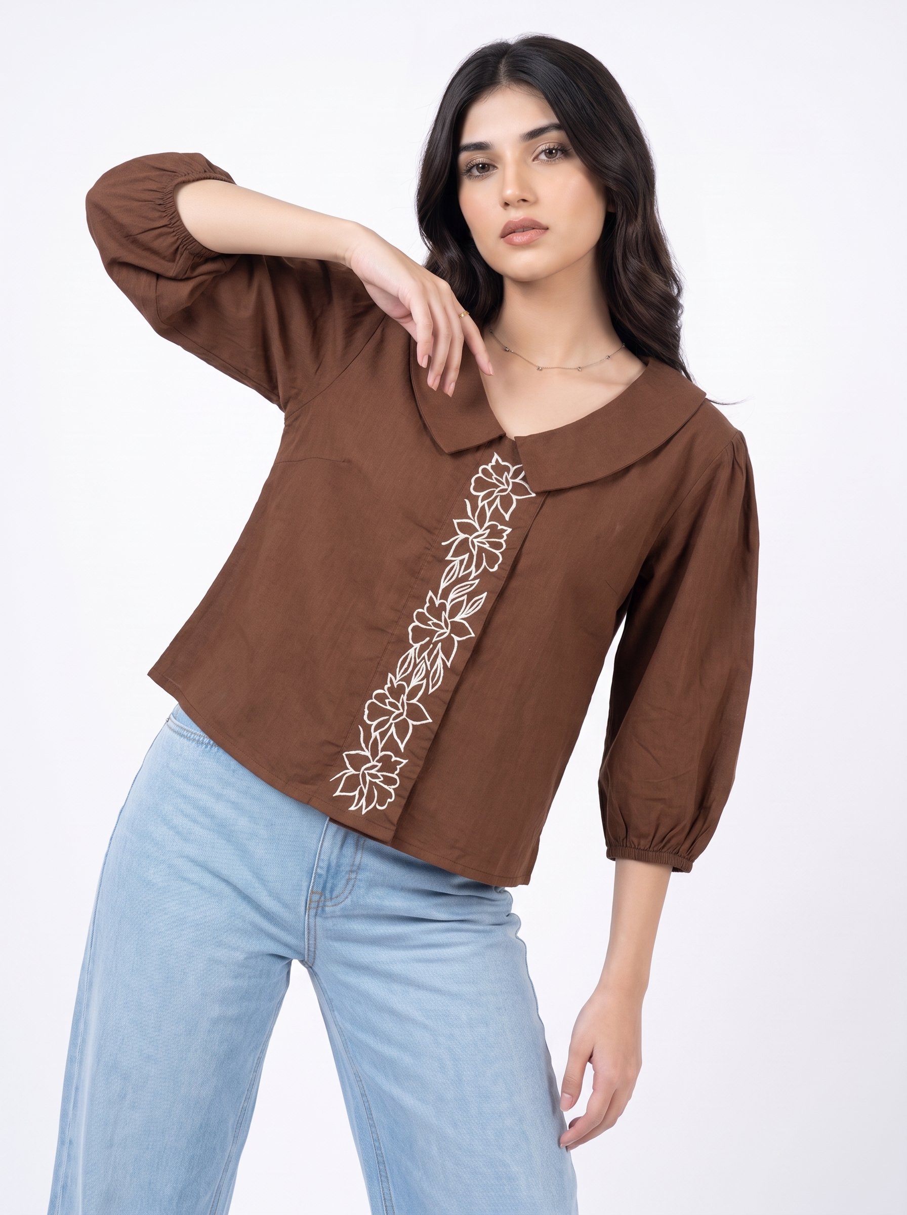 A woman modeling a brown cropped blouse with three-quarter puff sleeves, a wide collar, and white floral embroidery down the front, paired with light blue jeans against a plain white background.