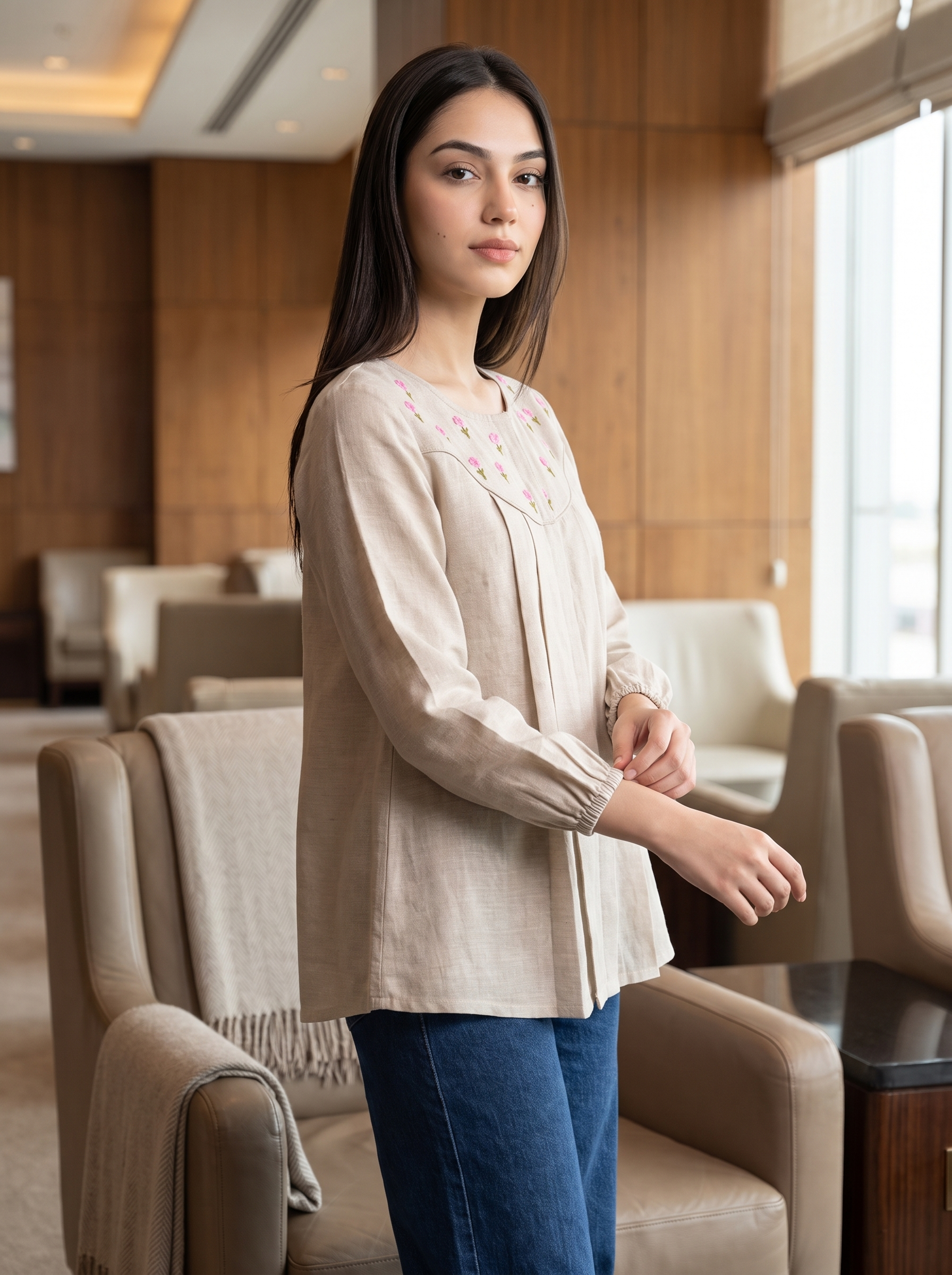 A woman wearing a beige long-sleeve linen blouse with pink and green floral embroidery on the chest, paired with blue jeans, standing in a modern lounge setting with beige armchairs.