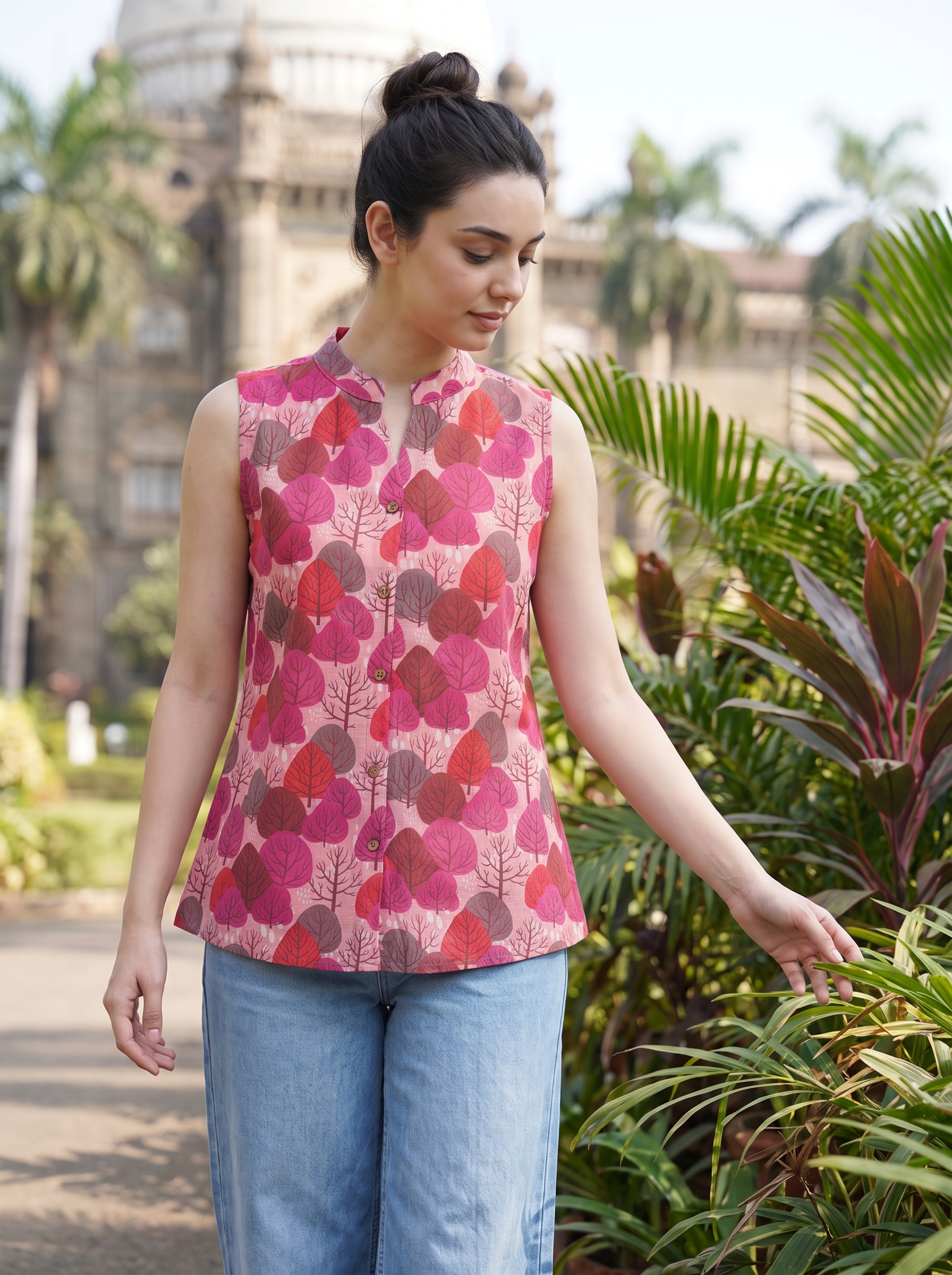 A woman wearing a sleeveless pink top with a red and brown leaf pattern, paired with light blue jeans, standing outdoors in front of tropical plants and a building.