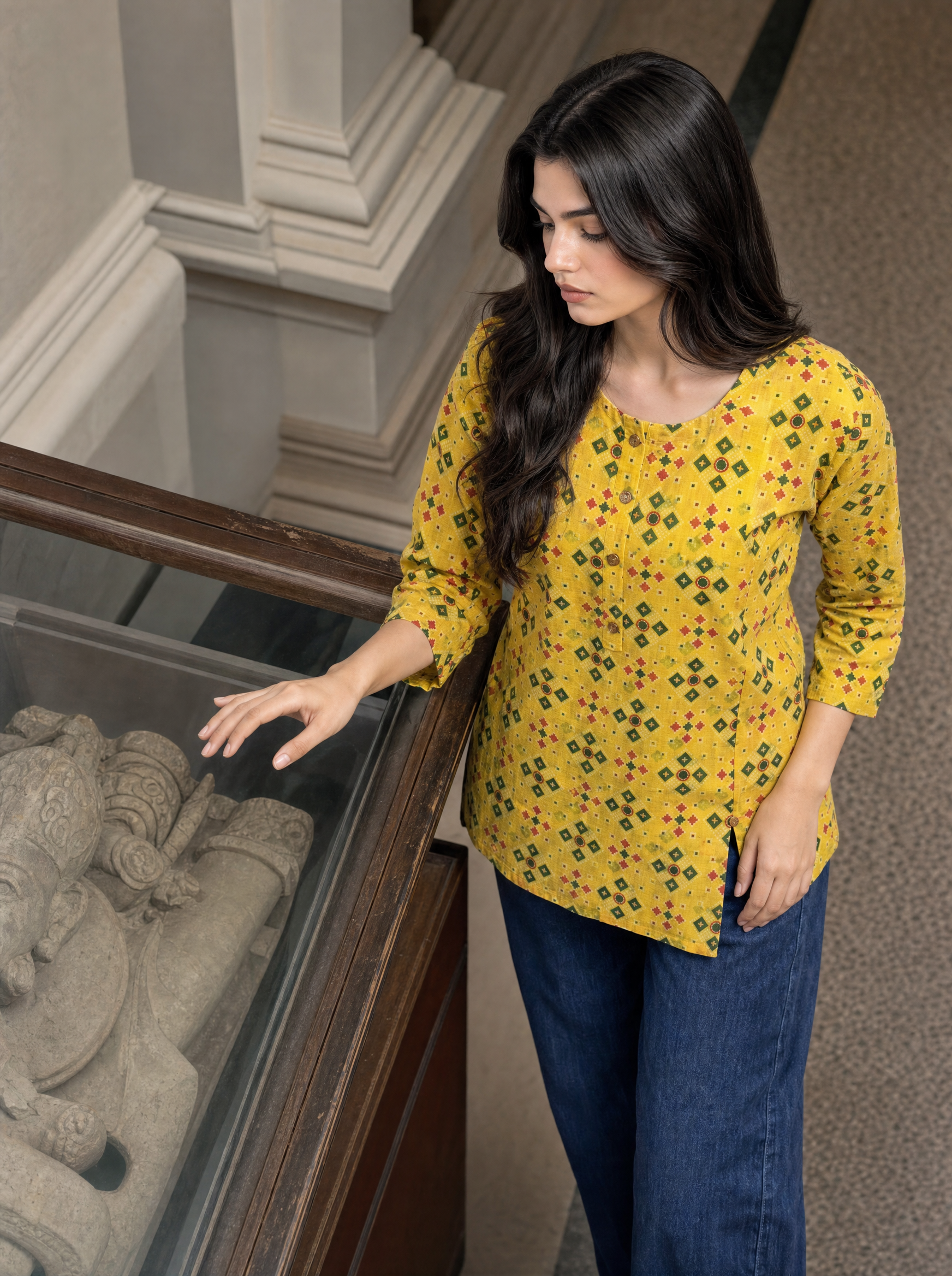 A woman with long dark hair stands next to a glass display case in a museum or gallery, wearing a yellow cotton kurta with a green and orange geometric pattern and dark blue jeans.