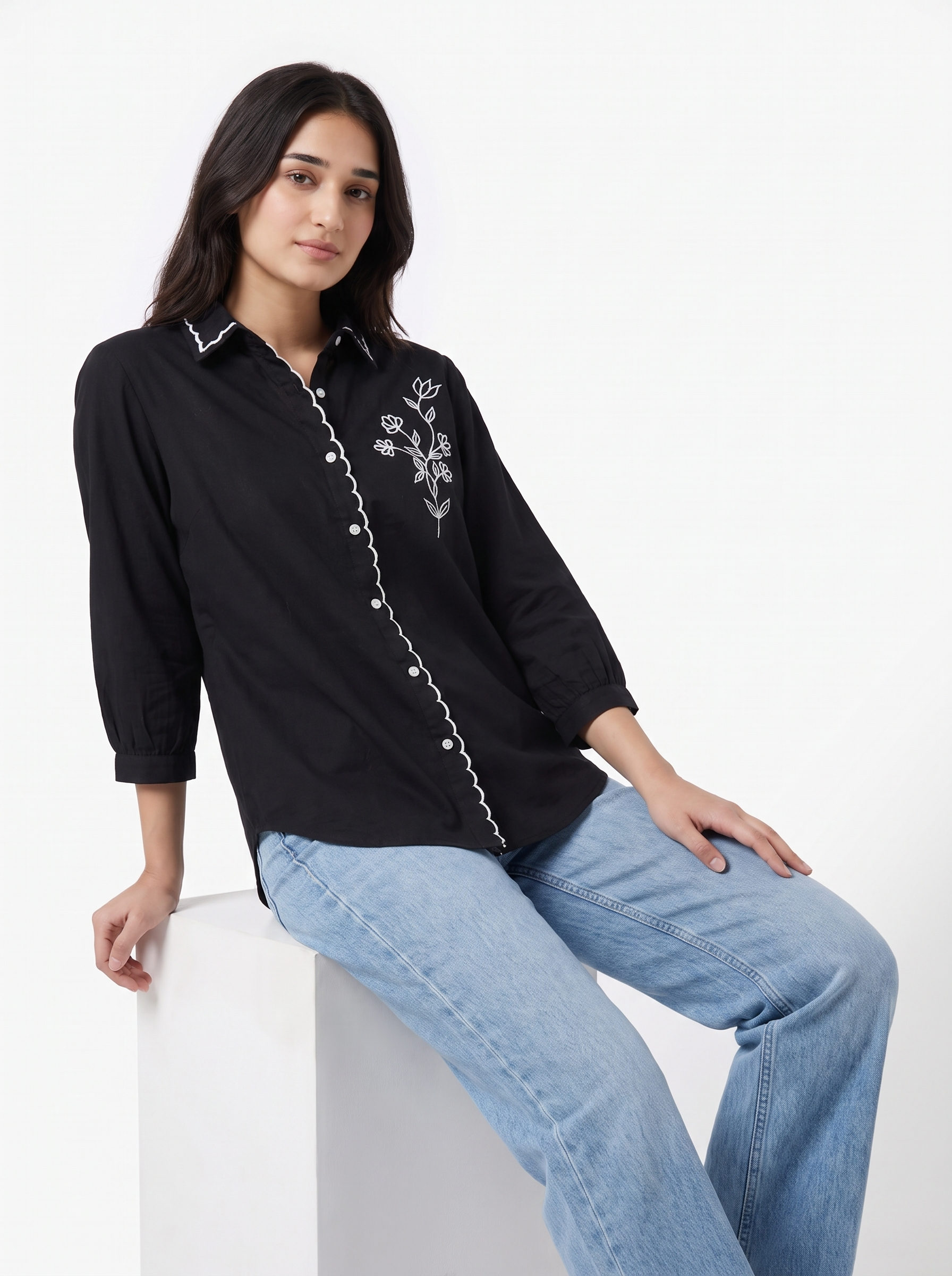 A woman wearing a black long-sleeve button-up shirt with white floral embroidery on the chest and light blue jeans, seated on a white block against a plain white background.