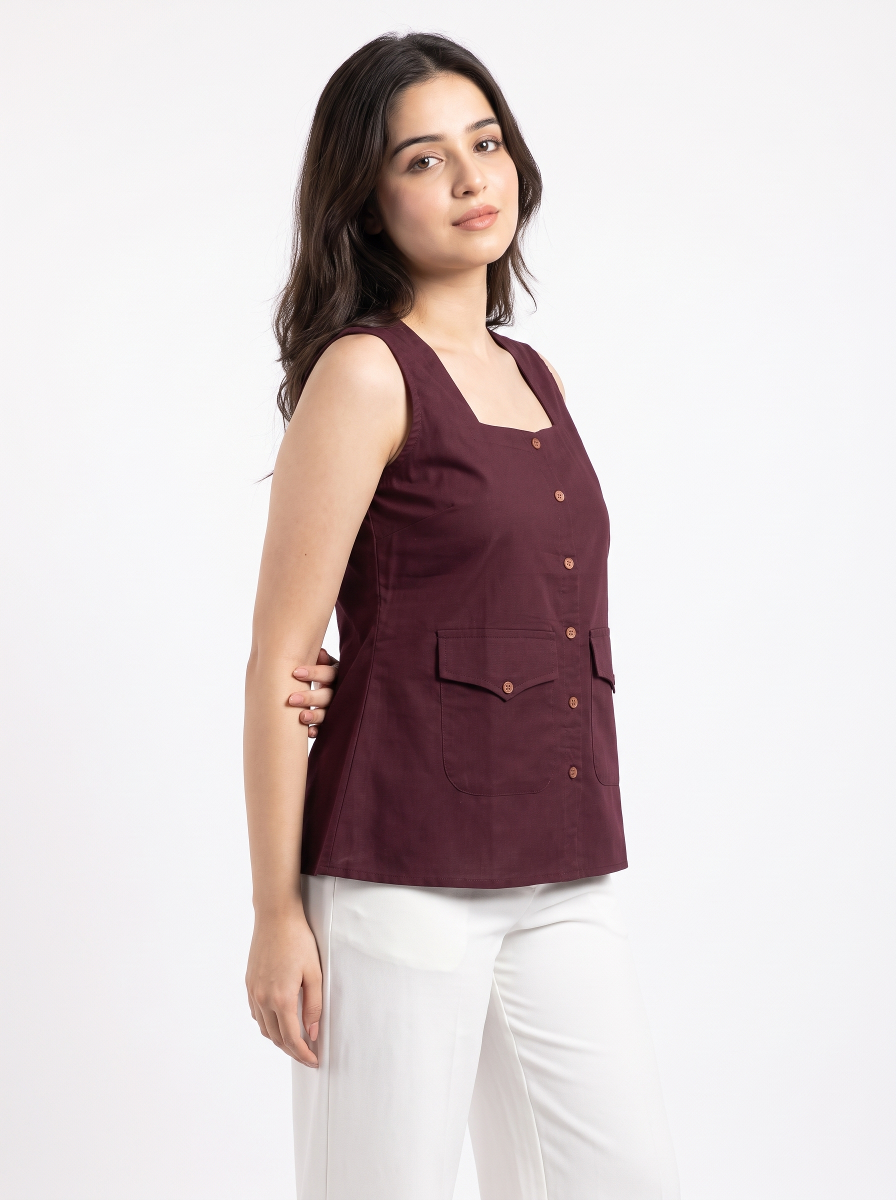 A woman modeling a sleeveless burgundy top with a square neckline, front button closure, and two front flap pockets, paired with white pants against a plain white studio background.