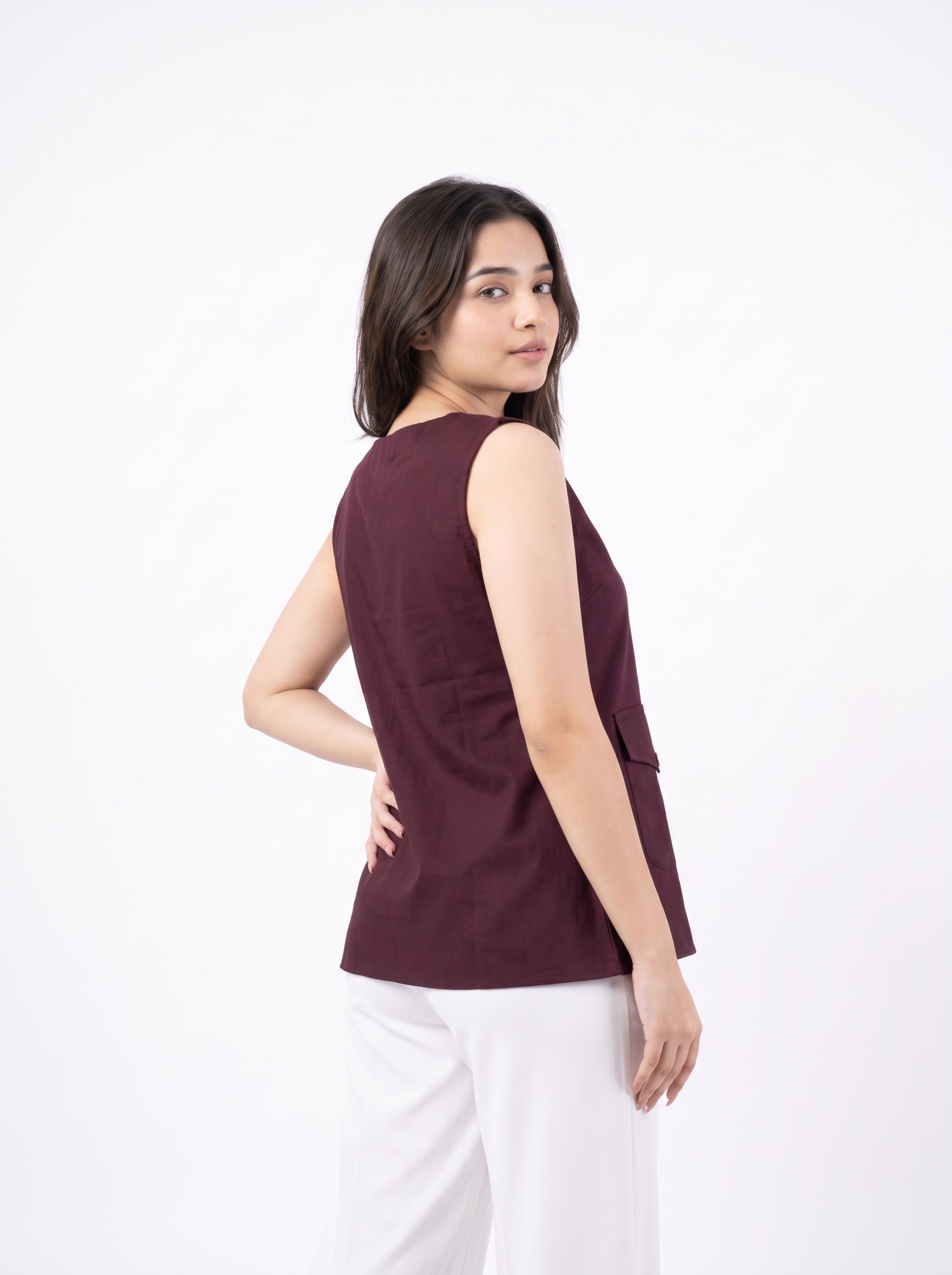 A woman modeling a sleeveless, boat-neck top in deep burgundy, paired with white trousers, photographed against a seamless white studio background.
