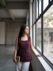 A woman stands by a large window in a modern industrial interior, wearing a sleeveless deep plum-purple top with front buttons and white pants, holding a small book or notebook.