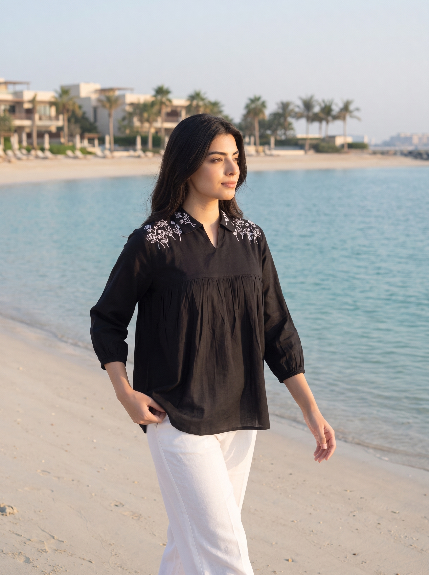A woman walking on a sandy beach wearing a black long-sleeve blouse with white floral embroidery on the shoulders and white pants, with the blue sea and resort buildings in the background.