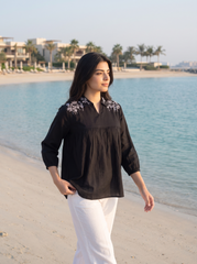 A woman walking on a sandy beach wearing a black long-sleeve blouse with white floral embroidery on the shoulders and white pants, with the blue sea and resort buildings in the background.
