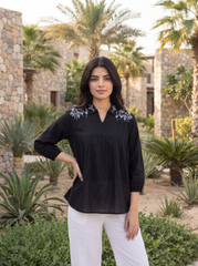 A woman stands outdoors in front of stone buildings and palm trees, wearing a black long-sleeve blouse with white floral embroidery at the collar and white pants.