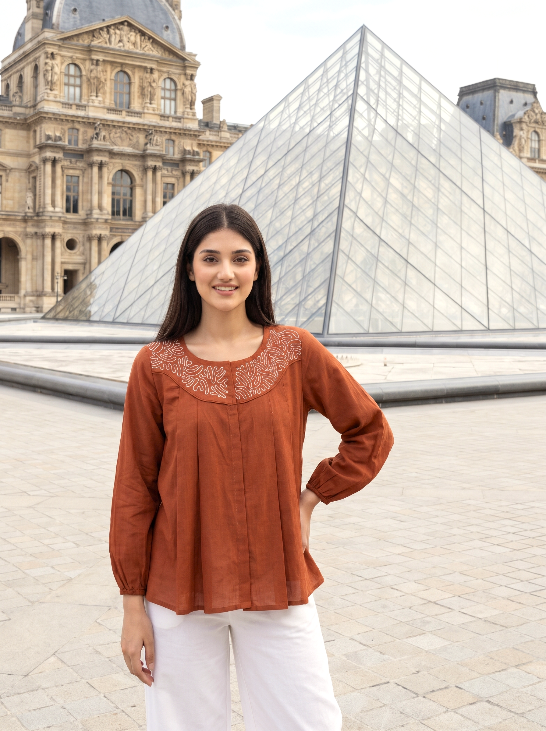 A woman stands in front of the Louvre Pyramid wearing a rust-orange long-sleeve blouse with white embroidery around the neckline, paired with white pants.