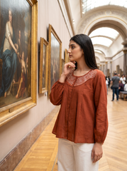 A woman in a rust-orange embroidered blouse and white trousers stands in a grand art gallery, looking at a large classical painting.