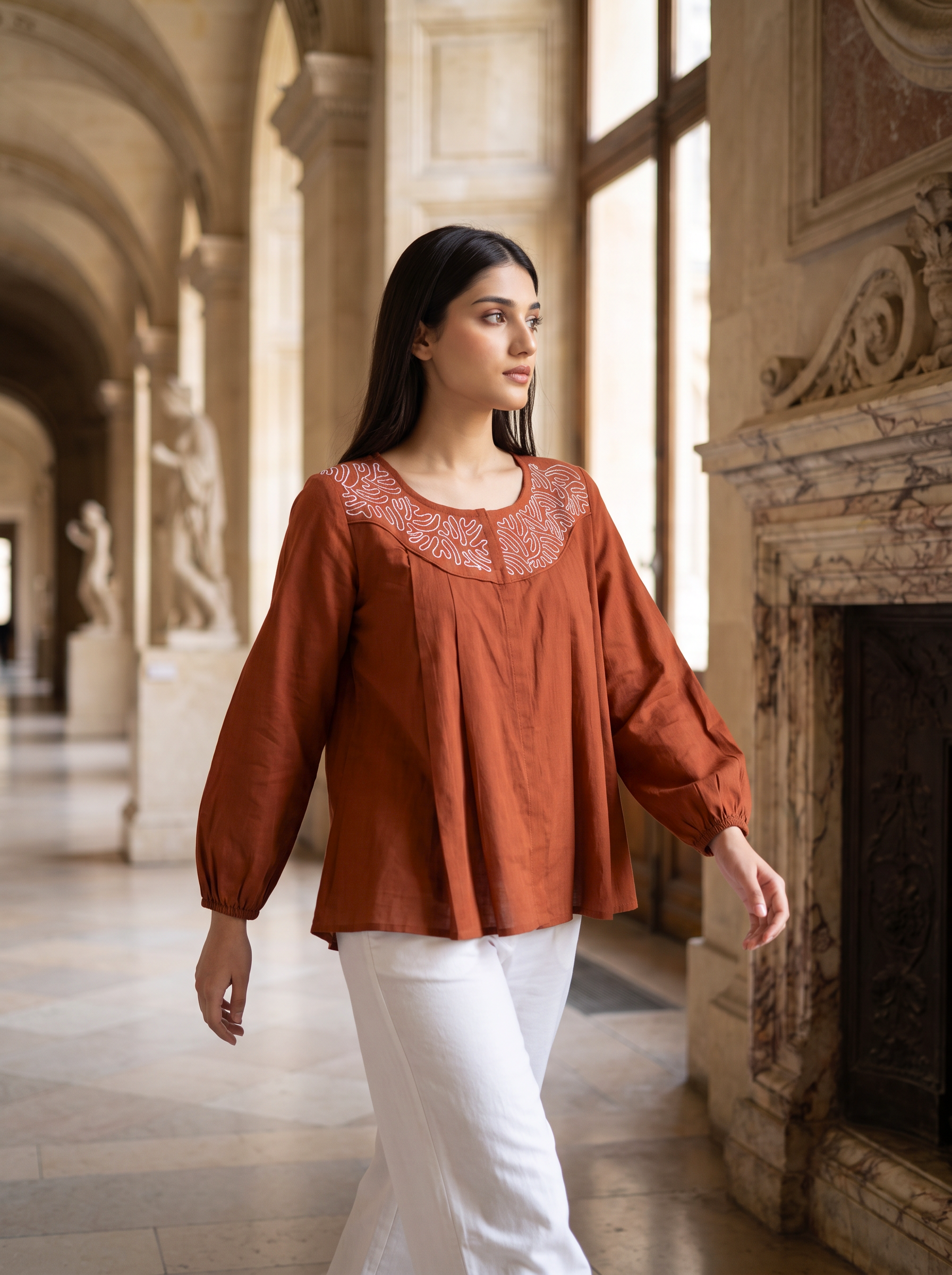 A woman wearing a rust-orange long-sleeve blouse with white pants walks through a grand, sunlit stone hallway with arches and a fireplace.