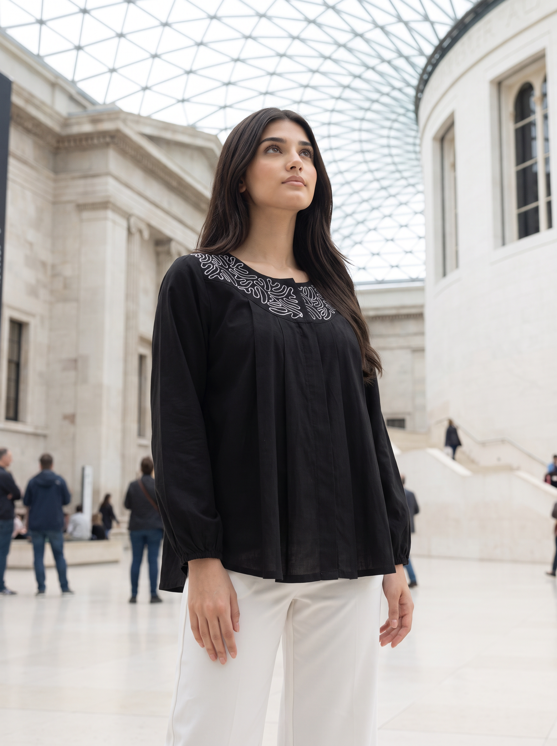 A woman wearing a black long-sleeve blouse with intricate silver embroidery around the neckline, paired with white trousers, standing in a grand, sunlit stone hall with a glass-domed roof.