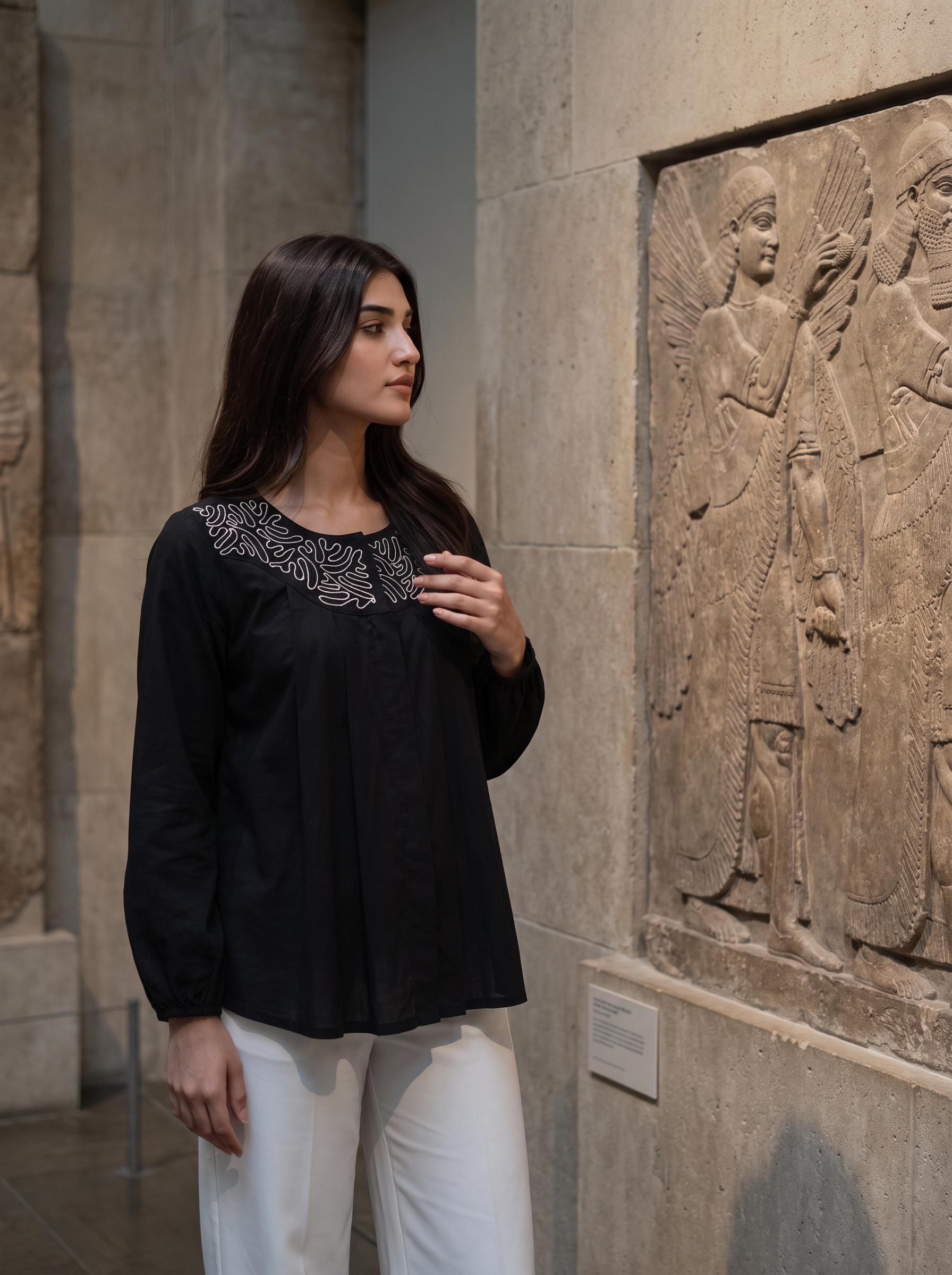 A woman wearing a black long-sleeve top with silver embroidery around the neckline and white pants, standing in a museum gallery next to a large stone relief carving.