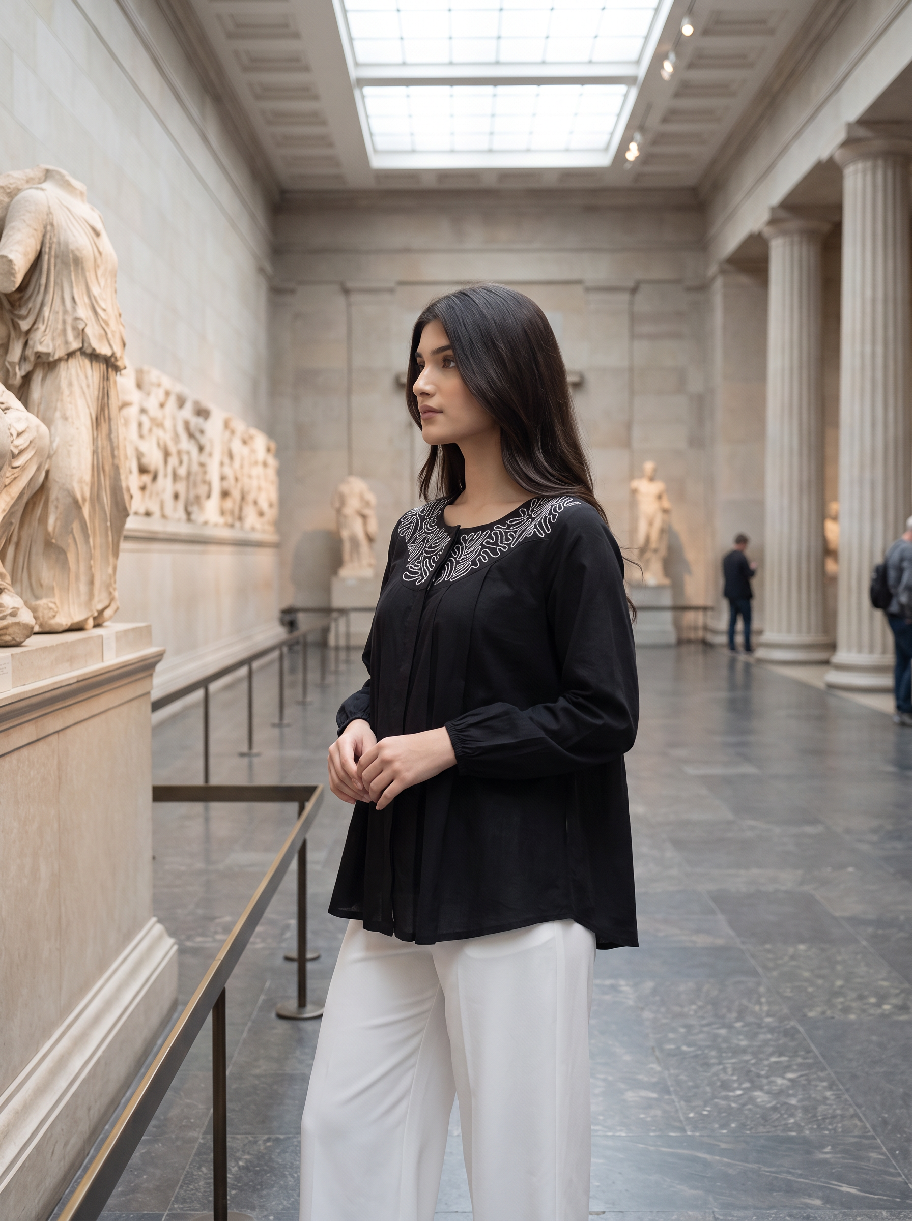 A woman wearing a black long-sleeve blouse with silver embroidery on the chest and white trousers, standing in a grand museum hall with classical statues and columns.