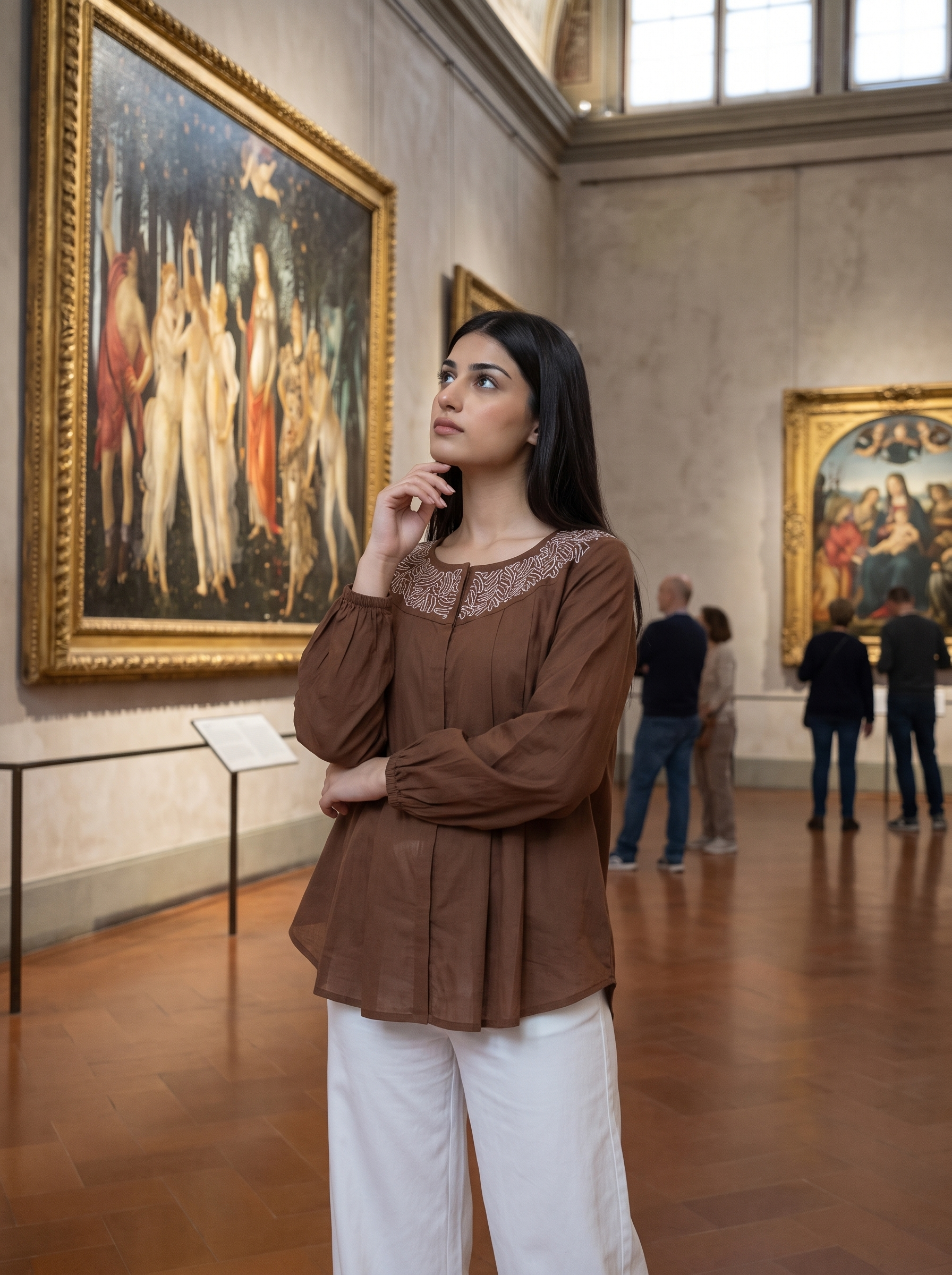 A woman in a brown long-sleeve blouse with white pants stands in a museum gallery, looking thoughtfully at a large classical painting on the wall.